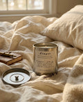 Linger soy-coconut candle in a brushed-silver tin, lit on cream linen bedding with a journal, pen and lavender — Ironic Paradoxists